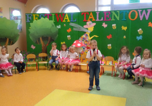 Chłopiec trzyma w ręku mikrofon, recytuje wiersz. Wokół chłopca siedzą dzieci.